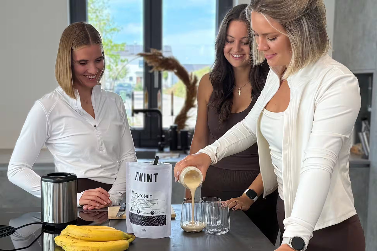 Drei Freundinnen in Sport-Outfits haben einen Kwint Bananen Shake gemixt und schenken ihn gerade in schöne Gläser ein