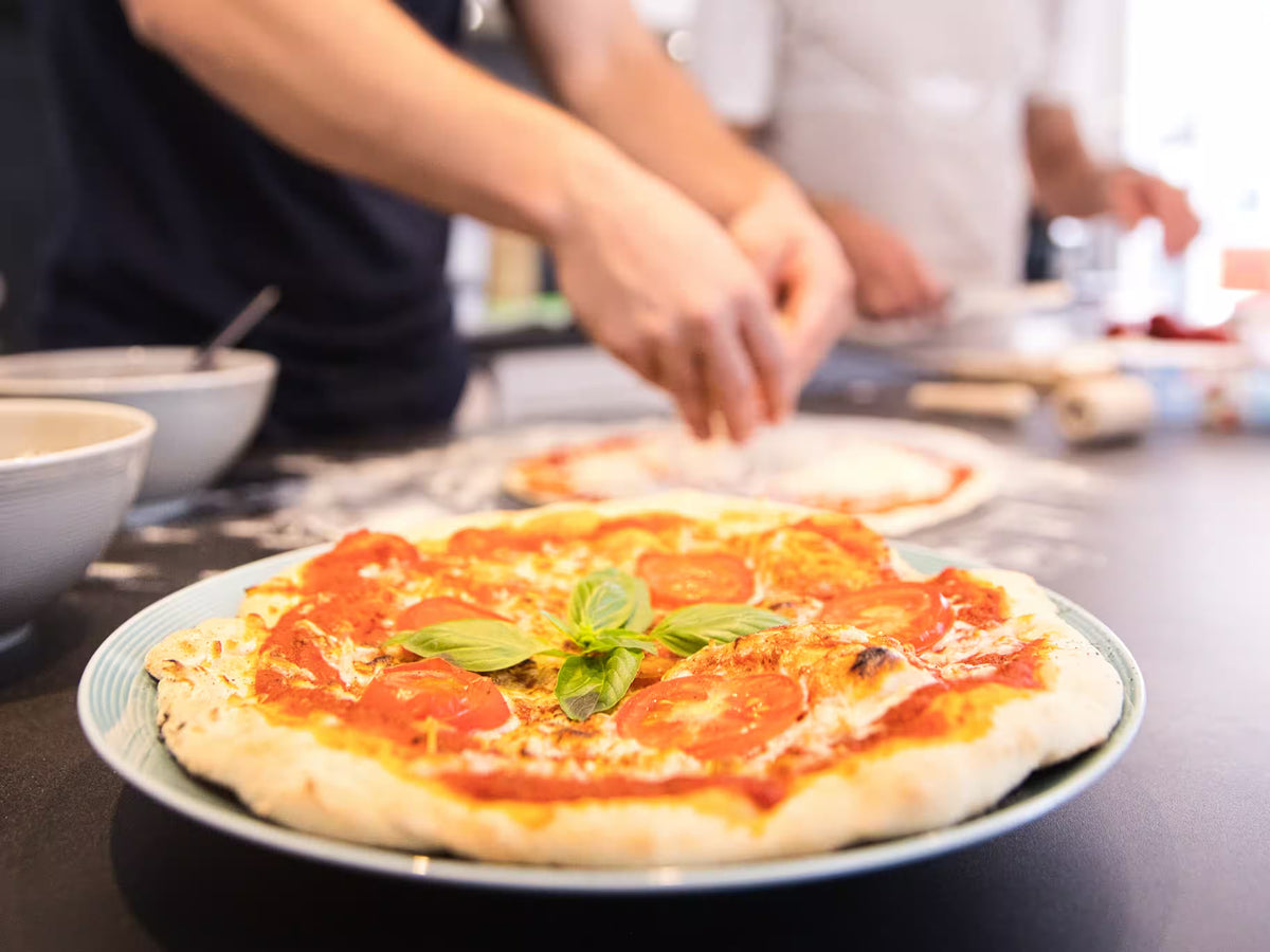 Gründer von Kwint backen zusammen eine Kwint Pizza mit Mykoprotein. Im Vordergrund ist eine fertige Pizza. Im Hintergrund belegen sie eine Pizza.